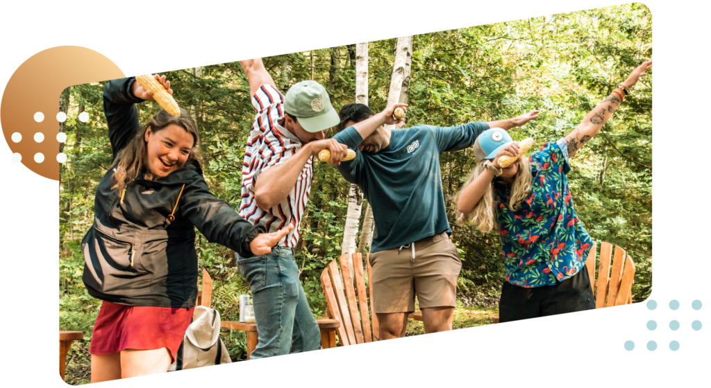 Four iBec team members holding ears of corn while dabbing. They are standing in a lush green forest with adirondak chairs behind them