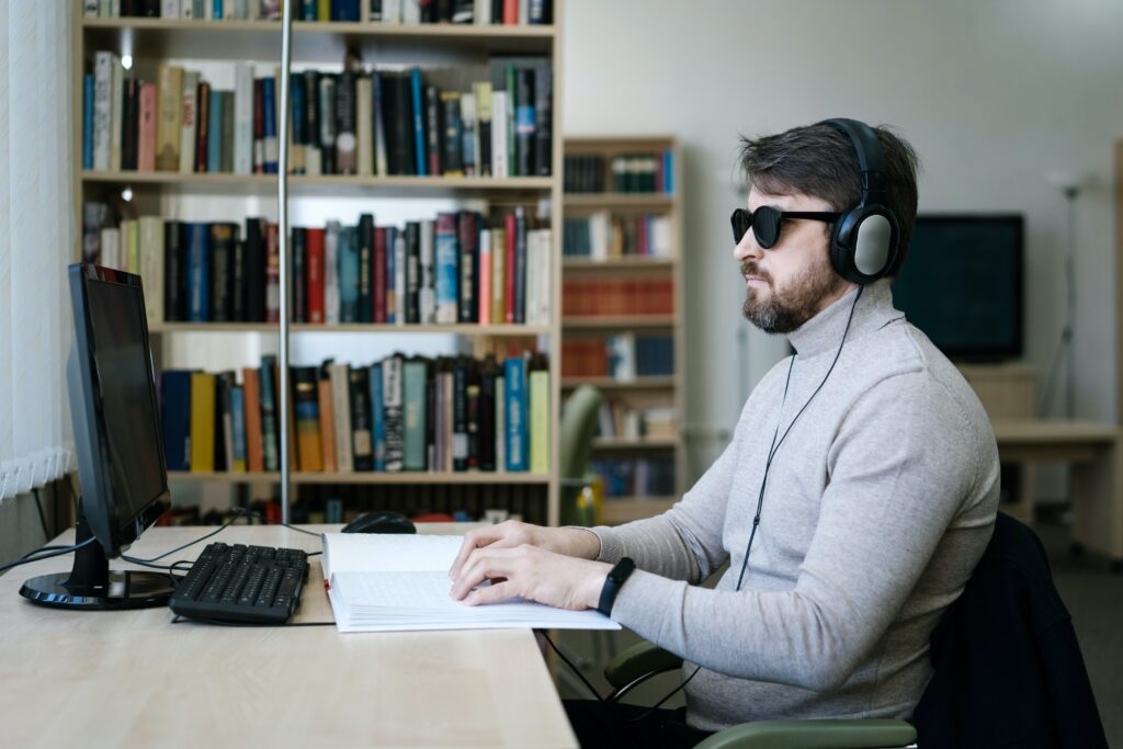 man wearing sunglasses sitting at a computer reading braille with headphones on in a library